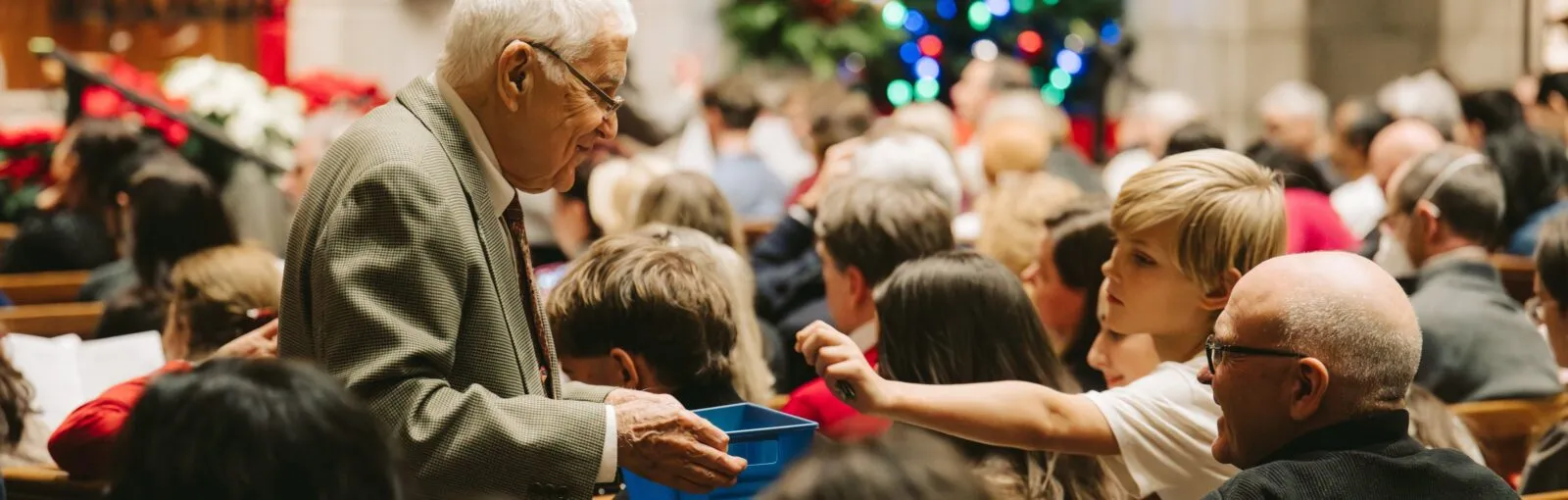 An usher holds out the offering basket as a child reaches in to give, at Carols by Candlelight.